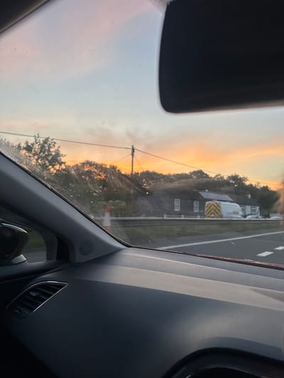 An outdoor, eye-level, close-up shot captures the view from inside a vehicle on a road at dusk. The sky exhibits a gradient of colors, transitioning from pale pinks and oranges near the horizon to a soft blue higher up, indicative of sunset. Silhouetted trees and power lines are visible against the colorful sky. In the mid-ground, a row of dark buildings with white-framed windows is situated behind a metal barrier. A white van with reflective chevron markings is parked in front of one of the buildings. The foreground is dominated by the interior of the car, including the dashboard, a speaker grille, and the car's side pillar, with the windshield showing some water spots. The road surface, marked with white lines, is visible in the lower right quadrant. The scene appears to be located in the Borough of Swale, United Kingdom.