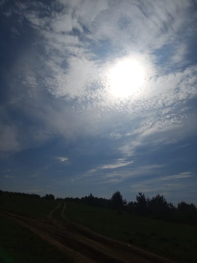The image depicts a rural landscape, likely in Ural'skoe, Russia, based on the provided location. A dirt road traverses a grassy hillside in the foreground, leading towards a tree line in the middle ground. The sky is predominantly blue with scattered clouds and a bright sun. The lighting suggests midday. There are no visible people, subjects, objects, actions, or interactions beyond the natural elements and the road's presence. The environment is characterized by natural terrain, vegetation, and atmospheric conditions.
