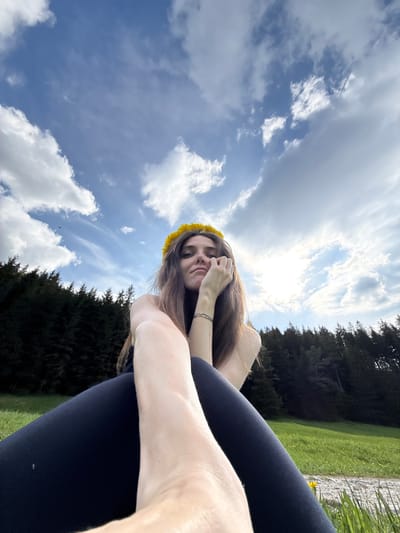 A young woman is captured resting outdoors under a bright blue sky with scattered white clouds, likely in Zakopane, Poland. The image is oriented diagonally, suggesting a low-angle perspective looking upwards. She has long, dark brown hair and is adorned with a vibrant yellow dandelion flower crown. Her face is directed towards the camera with a serene or contemplative expression, her eyes partially closed. One arm is gently raised, with her hand near her face. She is wearing dark leggings.
The background consists of a lush green landscape with abundant grass and a dense line of dark evergreen trees, characteristic of a forested or mountainous region. The sunlight is bright, indicating it is daytime. The scene evokes a sense of peace and connection with nature, possibly during a relaxing moment in a meadow or park. There is no visible text in the media file.