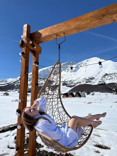 The image depicts an outdoor winter scene in Virak, Montenegro, under a clear blue sky. A female individual, dressed in a light-colored bathrobe, is reclining in a brown wicker-style hanging egg chair. Her long, light brown hair is visible, and her right hand rests near her forehead; her bare legs and feet are exposed. The chair is suspended from a rustic wooden frame, which is adorned with a string of white bulb lights. The ground is covered in extensive snow. In the mid-distance, several dark-roofed wooden cabins are scattered across the snow-covered landscape. The background features prominent snow-covered mountains with some visible patches of dark vegetation, beneath a sky displaying a faint contrail.