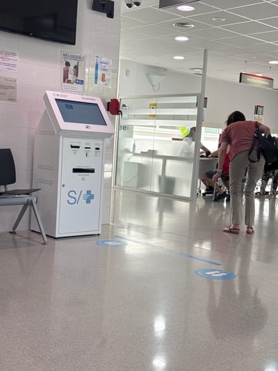 The image depicts the interior of what appears to be a public service office or healthcare facility. In the foreground, a white self-service kiosk with a touchscreen display is positioned to the left. The kiosk has various labels and symbols, including "S/+" and "Institut Català de la Salut." To the left of the kiosk is a grey metal chair with a black seat. The floor is a speckled grey material with a tiled pattern, and blue social distancing markers are visible on the floor, including a circular decal with two footprint icons.
In the middle ground, a glass partition separates the area with the kiosk from another section of the building. Through the glass, a desk is visible with individuals behind it, suggesting a service counter. To the right, a woman wearing a pink t-shirt and beige pants is bent over, interacting with something on the floor. She has a dark bag slung over her shoulder. Further to the right, other individuals are partially visible. A sign with the text "Atenció a la Ciutadania" (Citizen Attention) is mounted on the wall in the background. The environment appears to be well-lit with artificial lighting. The location is identified as L'Hospitalet de Llobregat, Spain.