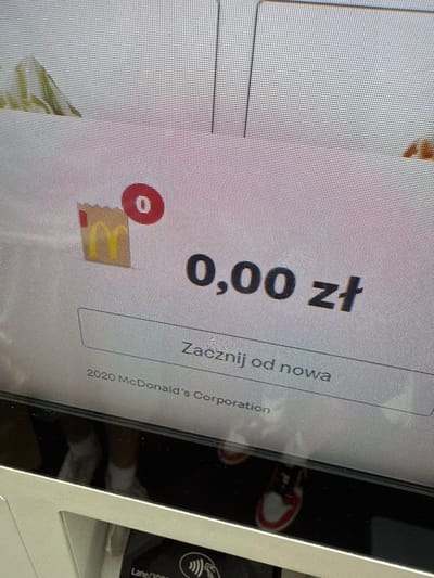 A digital display shows a McDonald's order summary. The total cost is displayed as "0,00 zł" (zero Polish zloty). Above this, a shopping bag icon with the McDonald's logo has a red circle with a "0" inside, indicating zero items in the order. Below the price, the text "Zacznij od nowa" (Start again) is visible. At the bottom of the screen, copyright information reads "2020 McDonald's Corporation". The display is part of a self-service kiosk, likely located in a McDonald's restaurant in Białystok, Poland. The reflection in the screen shows the lower legs and feet of two individuals wearing white socks and sneakers. One pair of sneakers is predominantly white with red accents, while the other pair is less discernible. The lighting appears to be artificial and overhead.