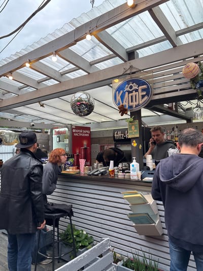 The image shows an outdoor cafe with a covered seating area. There are several people at the cafe. One man is standing behind the counter, talking on his phone. Another man is sitting at the counter, looking at his phone. A woman with red hair is standing at the counter, and a man in a black leather jacket is standing next to her. The cafe has a disco ball hanging from the ceiling, and there are bottles of alcohol behind the counter. The cafe appears to be located in the city of Komarovo, Russia, as indicated by the sign that says "КАФЕ" (Cafe).