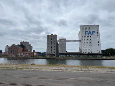 The image shows an industrial waterfront area in Odense, Denmark, under a cloudy sky. The foreground features a grey road with railway tracks. The middle ground is dominated by a body of water, likely a canal or harbor, with a paved embankment along its edge, showing some patches of grass and weeds.

On the far bank of the water, several large, imposing industrial buildings are visible. To the left, there is a multi-story brick building with a distinctive gabled roof, exhibiting signs of age and some graffiti. Adjacent to it are taller, more modern-looking concrete structures, including a prominent white silo-like building on the right, bearing the large blue text "FAF" on its facade. A connecting walkway bridges two of these concrete structures. The buildings show signs of disuse and graffiti, suggesting an industrial or post-industrial landscape.

The environment appears somewhat weathered, with overcast conditions contributing to a subdued and possibly melancholic atmosphere. The overall impression is one of a functional, perhaps historical, industrial site situated along a waterway.