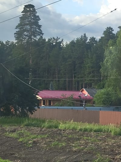 The image shows a rural scene in Chernaya, Russia. In the foreground, there is a field with dark soil and patchy green grass. A fence made of vertical wooden planks, some light brown and some reddish-brown, runs across the middle ground. Behind the fence, a wooden house with a red tiled roof is visible. Adjacent to this house is another structure with an orange roof. A dense forest of pine trees forms the background, with a tall pine tree prominent on the left side of the image. Overhead power lines are strung across the sky. The sky is partly cloudy with patches of blue.