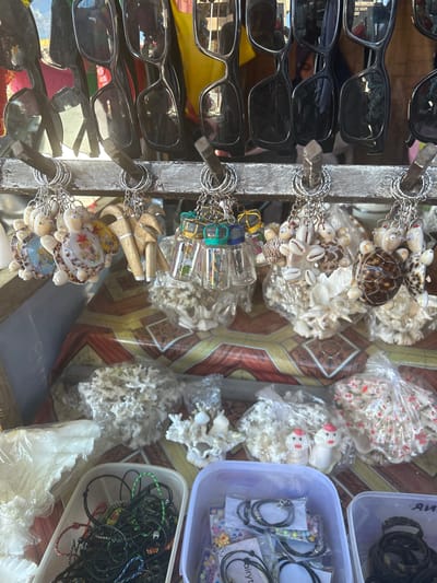 This is a close-up, eye-level shot of a street stall in Bira, Indonesia, displaying various souvenirs. The stall features a row of sunglasses hanging from hooks at the top. Below the sunglasses, a variety of keychains are displayed on a wooden bar. These keychains include small turtle figurines made of shells, wooden totems, and tiny glass bottles filled with what appear to be sand or shells. The background reveals more merchandise, including decorative shells, corals, and various bracelets or necklaces in plastic containers. The overall impression is one of a tourist-oriented market stall.