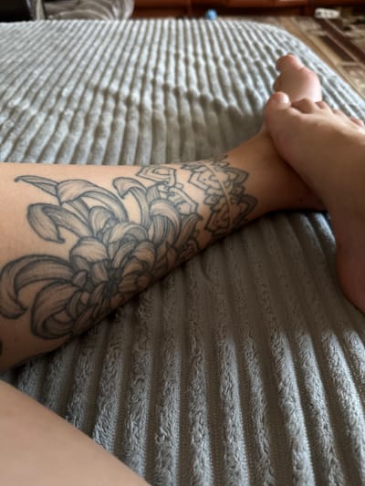 The image shows a close-up view of a person's legs and feet, resting on a textured gray blanket. A tattoo of a large flower, possibly a chrysanthemum, covers the left leg. To the right of the flower tattoo, there is a smaller, geometric mandala tattoo. The feet are bare and positioned slightly overlapping each other. The background is out of focus, showing what appears to be a carpeted floor and furniture. The scene is indoors, likely in a residential setting. No specific location in Piatykhatky, Ukraine, can be identified from the image.