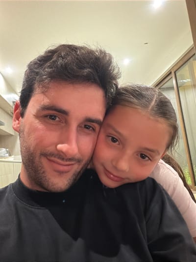 A man with dark curly hair and a beard smiles into the camera. A young girl rests her head on top of his. She also smiles at the camera. The man wears a black shirt. The girl appears to be wearing a white shirt and black sweater. The picture appears to be taken indoors with light-colored walls. There are multiple overhead lights.