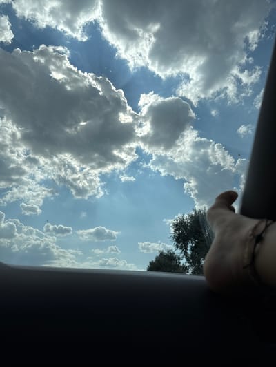 This media file shows a view of the sky through an opening, possibly a sunroof or window of a vehicle. The sky is filled with a mix of white and gray clouds against a bright blue backdrop. Sunbeams are visible, peeking through gaps in the clouds, suggesting it's a sunny day. On the right side of the frame, a person's hand and arm are visible, resting near the edge of the opening. The hand appears to be lightly holding onto something out of frame. Silhouetted against the sky are the tops of trees, indicating an outdoor, possibly rural or suburban, setting. The perspective is from below, looking upwards. The time of day appears to be daytime, with bright sunlight. The overall impression is one of a peaceful, perhaps travel-related moment, looking up at the sky. There is no visible text in the image.