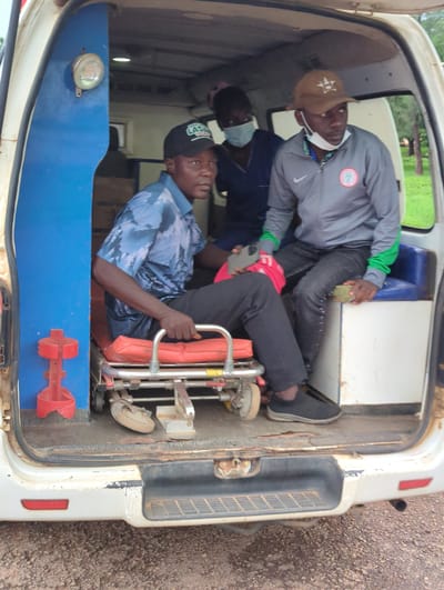 Three men are inside the rear of a white van. The man in the foreground wears a blue patterned shirt and a black baseball cap. He is interacting with a folding stretcher. Another man, wearing a gray sweatshirt with a Nigerian flag patch, is seated and appears to be looking at a phone. A third man is partially visible behind the others. The interior of the van is worn. The location is Gidan Waya, Nigeria, based on the cap on the man in the foreground that has the word LADA on it, a Nigerian NGO known to be active in that area.