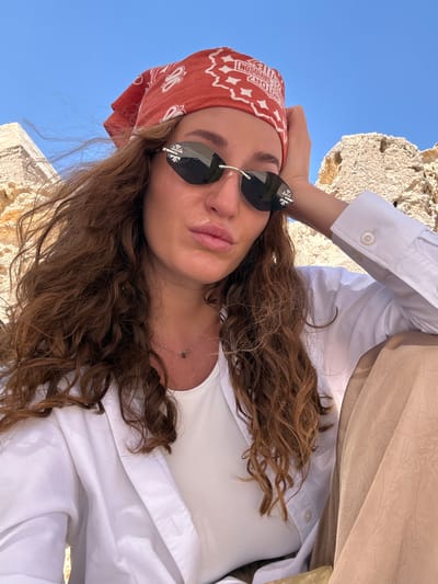 A woman wearing a red bandana and sunglasses is posing for a selfie in front of the pyramids of Giza, Egypt. She is dressed in a white button-down shirt and has long, curly brown hair. The sky is blue and clear.