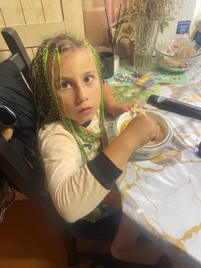 A young girl with braided hair interwoven with neon green strands sits at a table, holding a forkful of noodles from a bowl. She is wearing a light-colored long-sleeved shirt with black cuffs. The table is covered with a white and gold patterned tablecloth. To her left, there is a glass vase containing dried flowers, a bowl with sliced bread in plastic wrap, and various small objects. A black cylindrical object, possibly a flashlight or baton, lies on the table. In the background, wooden cabinets are visible. The scene appears to be indoors, possibly in a kitchen or dining area, with natural light contributing to the illumination. The girl's expression is one of slight surprise or curiosity.