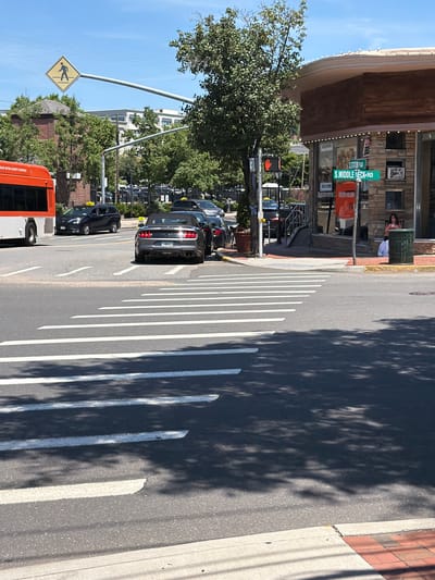 The image depicts a street scene in the Village of Great Neck Plaza, USA. The perspective is from a low angle, looking up at a street. The foreground shows a pedestrian crossing with white lines painted on the asphalt. To the left, a yellow diamond-shaped sign with a pedestrian symbol indicates a crosswalk.

In the background, several cars are parked or driving on the street. A dark-colored convertible is prominent, with its taillights illuminated. Further back, a row of other vehicles is visible. A bright orange trash bin with the word "FIRST AID" visible on it is next to a building with stone facade. Above the trash bin, a green street sign reads "S MIDDLE NECK RD". Another sign reads "STATION RD". A woman is visible sitting near the trash bin.

A large tree with green leaves partially shades the street and sidewalk. The sky is clear and blue, suggesting it is daytime with good weather. Shadows from trees are cast across the road, indicating sunlight. The overall impression is a typical sunny day in a suburban or small town environment. There is a bus visible on the far left of the frame.
