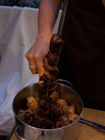In an indoor setting, a person's hand, adorned with a gold ring, is holding a skewer of grilled meat. The skewer is positioned over a metallic pot containing more pieces of meat. The background consists of draped fabric and a dark surface, suggesting a restaurant or home dining environment. The image does not provide any information about the city of Taganrog, Russia.