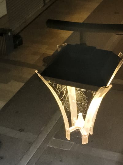 A close-up, low-angle shot captures a modern outdoor lamp post at night. The lamp is partially illuminated, casting a warm glow. The lamp shade is a dark, opaque rectangle. Below the shade, the lamp's structure consists of several curved, translucent white supports that converge towards the light source. A delicate, intricate spiderweb has been spun within the open structure of the lamp, catching the light and shimmering with dew or dust. The surrounding environment appears to be an urban setting, with a dark asphalt ground and hints of a building or wall in the background, suggesting it could be a public space like a street or park. The image does not provide specific identifiers for the city of Lagny-sur-Marne, France, beyond the user's provided context.