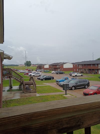 This image shows a residential area in Clarksville, United States of America. It looks out across rows of parked cars alongside low-rise brick apartment buildings with dark roofs. The sky is overcast. There are several exterior staircases and elevated walkways. A wooden railing is visible in the foreground. Grass and other vegetation provide a green element to the scene. The photo appears to be taken from an elevated position.