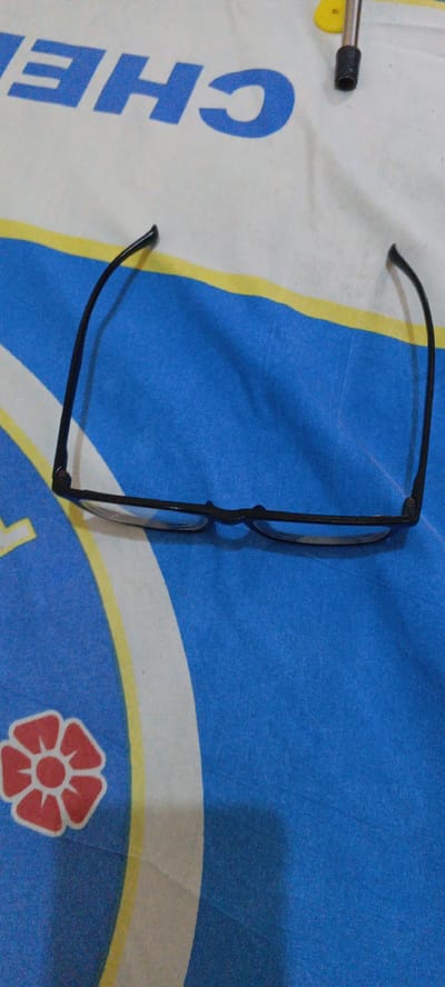 A pair of black-rimmed eyeglasses is positioned horizontally on a white and blue striped fabric, possibly a bedsheet or tablecloth. The fabric has a large, stylized red flower design and the word "CHE" printed in large blue letters. The eyeglasses are lying face down, with the temples extended. A yellow object and a black cylindrical object, possibly part of a pen or stylus, are visible in the upper right corner of the image. The lighting appears to be from above and slightly to the left, casting shadows from the eyeglasses onto the fabric. The image is a close-up shot, focusing on the eyeglasses and the immediate surrounding fabric. There is no indication of a specific location in Cirebon, Indonesia, beyond the possibility that the fabric's design might be culturally relevant.