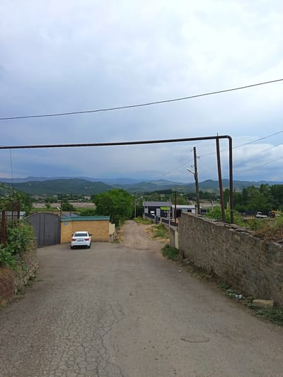 In a view looking down a paved road in Kasumkent, Russia, a white car is parked in front of a large gray gate. The road is cracked and shows signs of wear. To the left of the road is a stone wall, partially covered with vegetation. On the right side of the road, further down, a stone wall runs along the edge of the property. In the background, a range of green, rolling hills stretches across the horizon under a cloudy sky. There are a few buildings visible in the middle ground, including one with a green roof. Overhead, large metal pipes and wires are strung across the scene. No people are visible.