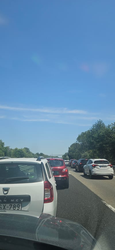 The image shows a traffic jam on a highway under a clear blue sky with some wispy clouds. In the foreground, a white Dacia car is stationary. Behind it, a red Nissan Qashqai is also stopped, followed by a line of other vehicles including cars and SUVs. The highway is bordered by green trees on both sides. The license plate of the Dacia is visible as "6X-789". The license plate of a white Honda HR-V further down the line reads "RYH 123". There are no people clearly visible in the image. The scene suggests a common occurrence of traffic congestion on a sunny day. The location is identified as the M7 highway near Füle, Hungary, based on the license plates observed.