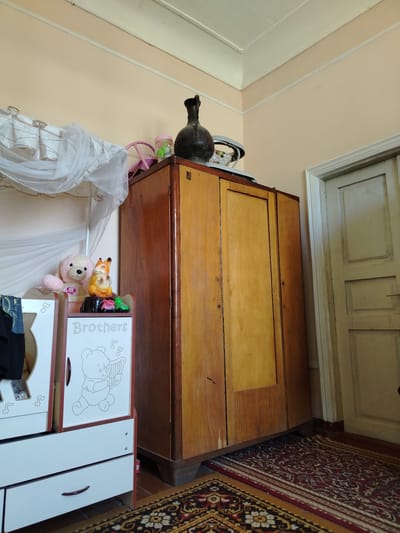This image depicts an interior scene, likely a bedroom or living area in a home in Usto Xo‘jamat, Uzbekistan. The primary objects are a large, wooden wardrobe standing to the right and a smaller white cabinet with drawers and a decorated door to the left. On top of the cabinet, there are several decorative items: a pink teddy bear, a figurine of a fox and a squirrel, and a small pink and green toy. A dark, tall vase or jug sits on top of the wardrobe. The room has pale yellow walls and a white ceiling with decorative trim. A door painted light beige is visible on the right side of the image. The floor is covered with patterned carpets in shades of red, black, and yellow. A white sheer curtain hangs from a rod above the white cabinet. Visible text on the white cabinet reads "Brothers" and includes a drawing of a bear playing a harp. The scene appears to be a still life, with no discernible activity or event taking place. The lighting suggests it could be daytime, but there are no specific cues about the weather or time of day. The overall impression is one of a domestic, lived-in space.
