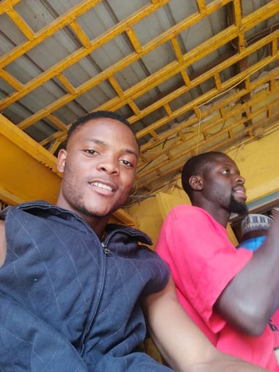 Two dark-skinned men pose for a selfie beneath a yellow-painted ceiling structure. The first man, in the foreground, smiles for the camera. He wears a dark, sleeveless, quilted jacket. Beside him, the second man, in a bright red shirt, glances off to the side with a neutral expression. A patterned white and blue cap peeks into the frame behind the second man. The overall lighting casts shadows.