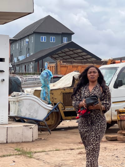 A woman with long, dark, wavy hair, wearing a leopard-print dress, walks towards the camera. She is holding a black clutch bag in her left hand. Behind her, there is a yellow Bomag compactor, a white truck, and an orange dump truck. In the background, a multi-story building with a dark gray facade and a dark gray roof can be seen. The scene appears to be an outdoor industrial area in Ojoto, Nigeria, with a dusty, unpaved ground. The sky is overcast with clouds.