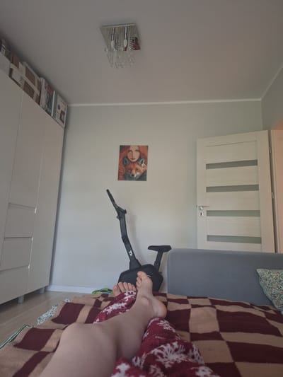 The image depicts an indoor scene, likely a bedroom, with a person's legs and feet visible in the foreground. The person is lying on a bed covered with a checkered blanket in shades of maroon and beige, and a red blanket with a white snowflake pattern. The room is sparsely furnished, with a light-colored door visible on the left, featuring vertical panels and a chrome handle. Below the door, on the wall, hangs a small, square painting of a woman with red hair embracing a fox. To the right of the painting, a black vacuum cleaner handle extends from the floor. Further to the right, a person's bare legs and feet are visible, suggesting they are lying down. A large, light grey piece of furniture, possibly a wardrobe or dresser, is visible on the right side of the image, with several boxes stacked on top of it. The walls are a neutral light color. The lighting suggests it is daytime. There is no visible text in the image. The location context is Mińsk Mazowiecki, Poland.