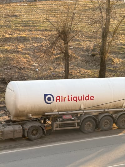 A white Air Liquide tanker truck is parked on a grey asphalt road in Kladovitsy, Russia. The truck's tires are visible on the right side of the image. The background features a field with dry grass and bare trees. Sunlight illuminates the scene.