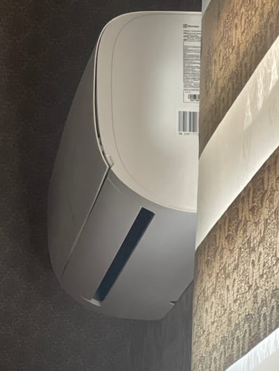 The image shows a close-up, vertical shot of a white air conditioner unit mounted on a wall. The air conditioner has a sleek, modern design with a curved front panel. A dark gray vent is visible on the lower part of the unit. To the right of the air conditioner is a window with blinds that have a textured, golden-brown pattern. The wall behind the air conditioner appears to have a dark gray, textured wallpaper. A label with "Electrolux" logo and some text in a small font is visible on the side of the air conditioner, along with a barcode and "SN 340F17773". There are no people or animals in the image. The scene appears to be indoors, likely in a residential or office setting. The image does not contain enough information to determine its location in the city of Atyrau, Kazakhstan.