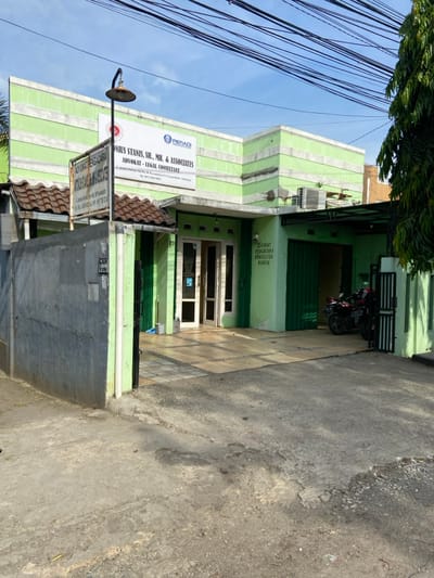 The image shows a green building with a sign that reads "Donies Stanisi, SH, MH, & Associates - Advokat - Legal Consultant" located at 99 Jalan Veteran, Purwakarta, Indonesia (latitude: -6.552, longitude: 107.437). The building has a tiled driveway leading to a double door entrance. There is a black motorcycle parked to the right of the building. The building has a green roof and a sign on the left wall that reads "JUICE PENGELOLA SAMPAH". In the background, there are several power lines and a clear sky. The surrounding area is a quiet residential street.