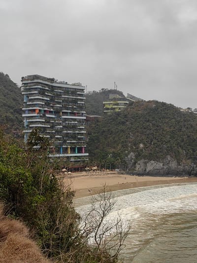 The image depicts a coastal scene in Hải Phòng, Vietnam. The foreground features a beach with light brown sand and waves breaking onto the shore. Several people are walking on the beach. The focal point is a tall, modern hotel building with multiple balconies and vertical gardens, some of which are painted in bright colors. The hotel is built into the side of a hill covered in dense green vegetation. Another building, smaller and lime green in color, sits atop another hill in the background. The sky is overcast and gray. Trees and shrubs in the immediate foreground obscure part of the view.