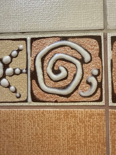 The image displays a close-up view of decorative tiles. The central tile features a brown border and a textured reddish-brown interior, overlaid with a white spiral design outlined in dark brown. To the left, a tan tile with a textured surface contains a pattern of white circular elements connected by thin lines. Above these tiles, a light-colored tile with a slightly raised, textured surface is visible. Below the decorated tiles, a larger, uniformly textured light brown tile forms the lower portion of the frame. The image appears to be taken in Taldybulak, Kazakhstan, suggesting these tiles might be part of a building's facade, interior wall, or a decorative element within the city. There are no people or actions depicted.