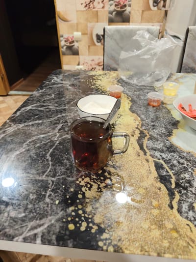 A close-up shot captures a dark marble countertop with a swirling gold and black pattern. On the countertop, a clear glass mug filled with dark tea and a spoon rests in the foreground. To the left of the mug, a white bowl contains a white powdery substance, likely sugar. Behind these items, several small plastic containers with colorful contents, possibly food coloring or spices, are scattered. Further back, a crinkled plastic bag appears to hold an unknown item. The background reveals a tiled wall with a floral design and hints of kitchen appliances. The lighting suggests an indoor setting, possibly during the day given the reflections on the countertop. No people are visible in the image. The overall impression is that of a kitchen or dining area where food preparation might be taking place.