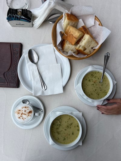 An overhead view captures a dining table set for a meal, likely in a restaurant or cafe in Šavnik, Montenegro. The table is covered with a light-colored, textured tablecloth.
The primary subjects are two bowls of soup, each served in a white bowl on a white saucer. The soup is light green or yellowish, garnished with chopped green herbs, possibly parsley, and appears to contain small vegetable pieces. A spoon rests in one of the soup bowls. To the left, a rustic woven basket holds several pieces of white bread, some with a golden-brown crust, lined with white paper napkins.
A person's hand, with dark-colored nail polish, is visible at the top right of the frame, resting near one of the soup bowls, suggesting the presence of a diner. In the lower right corner, a small white cup containing a hot beverage, likely a cappuccino or coffee, is topped with frothed milk and sprinkled with a brown powder, such as cinnamon or cocoa. It sits on a matching saucer with a small spoon.
Other items on the table include a stack of folded white paper napkins, a silver spoon resting on a white plate, and a clear glass, possibly an ashtray or beverage glass. A rectangular dispenser with various packets, possibly sugar or creamers, is also visible, bearing the brand name "MANUEL" on its side. A dark brown wallet or menu holder with an embossed pattern is positioned towards the bottom center. The scene suggests a casual meal for two.