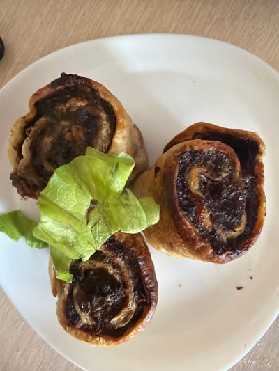 The image shows three rolled pastries on a white plate. The pastries appear to be filled with a dark, possibly sweet, filling. One pastry is stacked on top of another, and a third pastry is positioned to the left. A piece of green lettuce is placed in front of the pastry on the left. The plate is on a light-colored wooden surface. The lighting suggests it is daytime, and there are no external weather cues. The scene appears to be a casual setting, likely a kitchen or dining area. There is no visible text in the image.