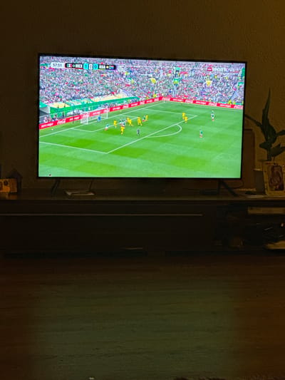 The image shows a television screen displaying a soccer match. The score shown on the screen is 57:51, with Mexico leading 1-0 against RSA. The stadium is filled with spectators, and there are various advertisements around the field, including Coca-Cola. The television is placed on a dark wooden cabinet. To the right of the television, there is a potted plant and a framed picture. The overall environment appears to be a living room or entertainment area. There is no information in the image to indicate the location in the city of Hüsen, Switzerland.