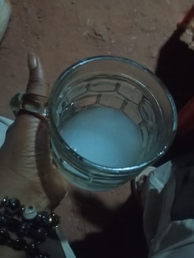 A hand holds a partially filled glass mug with a foamy white liquid. The mug is clear with a pattern of hexagonal recesses. The hand is dark-skinned and wears a beaded bracelet. The background is dark and grainy, revealing a reddish-brown surface and glimpses of clothing. The mug is the central focus, with the liquid suggesting it might be milk or a similar beverage.
