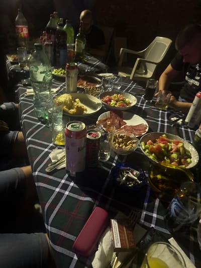 This image captures a casual gathering around a table laden with food and drinks. The scene appears to be outdoors, possibly in a backyard or patio setting, given the presence of chairs and the slightly uneven ground visible in the background. The table is covered with a dark green and white checkered tablecloth, which is characteristic of picnic or outdoor dining setups.
Several people are partially visible in the frame, with their hands and arms reaching for food or drinks, suggesting an ongoing meal or social interaction. There are various food items on the table, including a salad with tomatoes and cucumbers, sliced meats (likely salami or prosciutto), and a bowl of nuts, possibly almonds. Beverages are also abundant, with several bottles of water and other drinks, as well as cans of soda or beer, scattered across the table. Some glasses, both empty and partially filled, indicate that people are actively drinking.
A prominent detail is a cigarette pack with text in Bulgarian, which reads "TEREA. This tobacco product harms your health and leads to addiction." This suggests a location where smoking is present and possibly common.
The lighting suggests it might be late afternoon or early evening, as the overall scene is dimly lit, hinting at the approaching twilight or an indoor setting with ambient light. The mood seems relaxed and convivial, typical of a gathering of friends or family. The image is taken from a slightly elevated angle, looking down at the table, providing a comprehensive view of the spread and the activity.