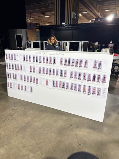 The image depicts an indoor, industrial-style environment, consistent with a large studio or warehouse, located in Issy-les-Moulineaux, France. In the foreground, a large, white rectangular board stands vertically, serving as a display. This board is covered with multiple small, rectangular print-outs, arranged in horizontal rows. Each print-out displays an image of a model wearing various lingerie or swimwear items, and many include handwritten notes or labels below the images. Centered at the top of the board, the phrase "— EVERYDAY SENSUALITY —" is written in dark capital letters.

Behind the board, a woman with dark hair, wearing a dark blue hooded jacket with a prominent fur-lined collar, is visible. She appears to be observing the contents of the board. Further back and to the right, a second individual with dark, curly hair is partially visible, positioned behind what appears to be a table or surface, and seems to be engaged with an object, possibly a laptop or tablet.

The background consists of dark staging structures, vertical wooden panels, and exposed industrial ceiling elements including light-colored beams and various fixtures. To the far left, a white door frame is visible against a dark wall. The floor is concrete, displaying visible cracks, stains, and general wear. A portion of a dark, round stool or chair is visible in the bottom right corner of the frame.