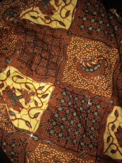 The image displays a close-up, overhead view of a textile fabric featuring a complex, multi-panel pattern. The primary colors observed are various shades of reddish-brown/maroon, golden-yellow, and black/dark brown. The pattern consists of interconnected sections: some panels display intricate geometric motifs, including repeating floral or star-like shapes within squares and diamonds, predominantly in dark brown on a reddish-brown background. Other panels exhibit curvilinear designs, such as stylized leaves, vines, and paisley-like elements in dark brown on a golden-yellow ground, with some accents of reddish-brown. The fabric surface appears to be slightly wrinkled or draped, and scattered specular highlights indicate a slight sheen or reflectiveness in localized areas. No individuals, specific objects beyond the fabric, actions, or direct environmental cues are visible. The image contains no visual elements to confirm a specific location within the city of Na Sang, Laos.