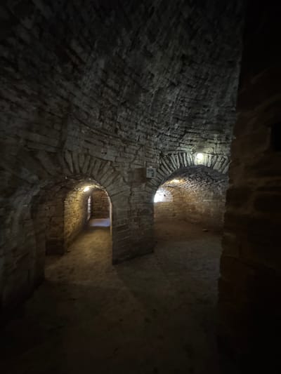 The image shows the interior of a dark, stone spiral staircase. The rough-hewn stone walls are illuminated by several artificial lights placed at intervals up the structure, casting an eerie glow. The staircase itself is made of brick, with the individual bricks visible, and it winds upwards and out of view. There appear to be openings or landings visible at different levels. The overall impression is one of an ancient and possibly abandoned structure, with a sense of depth and mystery. The scene is devoid of any people or clearly identifiable objects beyond the architectural elements. The time of day and weather are not discernible from the image. There is no visible text. The location context suggests this might be in Posyolok Imeni Morozova, Russia, but this cannot be confirmed from the image alone.