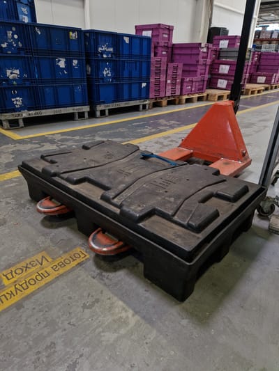 A black plastic pallet with orange fork extensions underneath is positioned on a concrete floor in a warehouse setting. Yellow stenciled text is visible on the floor in front of the pallet, with one line reading "ГОТОВА ПРОДУКЦИЯ" and another reading "ИЗХОД". Behind the pallet, a red and black manual pallet jack is partially visible, with "TOYOTA" text on the red portion. To the left, stacks of blue plastic storage bins are visible, and further back, stacks of purple plastic storage bins are present. The overall environment suggests an industrial or storage facility. The location is specified as Bahovitsa, Bulgaria, though no visual cues in the image confirm this specific location.
