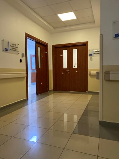 The image displays an empty, brightly lit hallway within a medical facility in Sidi Chahmi, Algeria. The walls are painted a light cream color, transitioning to a light yellow-beige wainscoting section below a horizontal beige handrail on both the left and right sides. A dark gray baseboard runs along the bottom of the walls. The floor is covered with highly reflective light beige square tiles, showing reflections of the overhead lighting, with a section of darker gray tiles on the right side of the hallway. The ceiling is a white suspended grid system with embedded rectangular fluorescent light fixtures, one of which is visibly illuminated in the upper center.

On the left side of the hallway, a dark brown wooden door stands partially open, revealing a continuation of the corridor with another brown door further down. Above the left handrail, a clear acrylic sign with white lettering is mounted, displaying "الأعصاب" (Neurology) and "العيادات المستعجلة" (Emergency Consultations) in Arabic, followed by "Neurologie" and "Consultations d'urgence" in French, with the room number A040. A small black square electrical fixture is positioned below this sign.

Centered in the image are closed double doors made of dark brown wood, each featuring two elongated vertical translucent glass panels. These doors are equipped with a silver-colored handle and lock mechanism. On the right wall, a partially visible clear acrylic sign indicates "صيدلية" (Pharmacy) in Arabic and "PHARMA" in French, accompanied by the room number A010. There are no individuals, active interactions, or movable objects present in the scene.