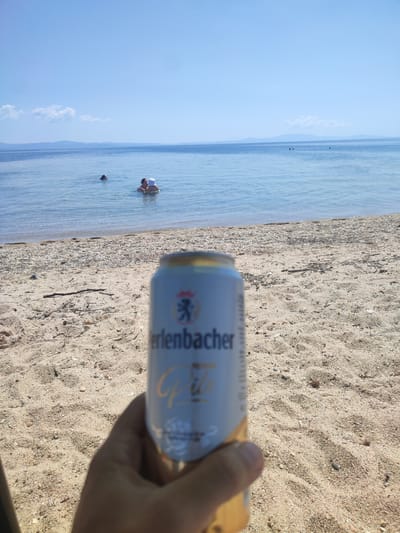 A first-person perspective shot shows a hand holding a can of Erlenbacher Pils beer on a sandy beach. In the shallow, clear blue water of Nea Potidea, Greece, two people are visible, one with a child. The horizon shows a faint outline of mountains under a bright blue sky with a few wispy clouds. The beach is composed of fine sand mixed with small pebbles and driftwood.