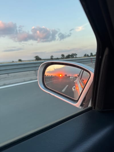 The image shows a view from inside a car on a highway, likely near Belozem, Bulgaria, at sunset. A side-view mirror reflects the setting sun, casting a warm orange glow across the sky and the road. Clouds are visible in the sky, tinged with pink and orange. The highway stretches into the distance, with other cars visible as blurred lights. Trees and vegetation line the sides of the road. The interior of the car, including the dashboard and a speaker, is partially visible in the foreground. The overall impression is one of a scenic drive during golden hour.