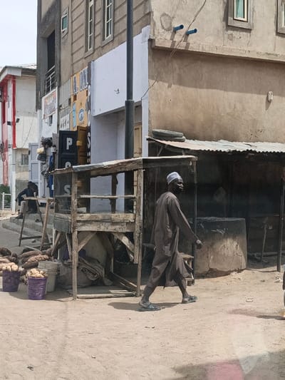 The image displays an outdoor street scene in Hotoro South, Nigeria, under bright daylight, with compacted dirt serving as the ground surface. In the foreground, a dark-skinned male subject, wearing a dark brown tunic, a white cap, and black sandals, is observed mid-stride, moving from right to left across the frame. To the left of the central path, two purple plastic baskets filled with white root vegetables are placed on the ground, accompanied by a pile of larger brown root vegetables. A two-tiered, wooden stall with mesh sides occupies the central midground. Behind this stall, to the right, stands a structure with a corrugated metal roof, topped by two stacked tires. Adjacent to this structure is a large, circular, concrete or earthen installation. In the background, a second male subject, dressed in dark clothing, is seated on wooden steps near a building on the left. The background consists of several multi-story buildings exhibiting varying architectural styles and finishes, along with a prominent black utility pole and overhead wires.