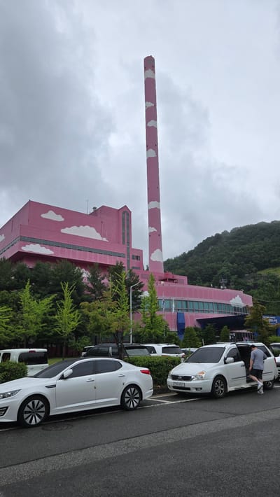The image shows the exterior of the Gwangmyeong City Energy Center, a power plant located in Gwangmyeong, South Korea. The building is painted bright pink and decorated with white cloud motifs. A tall, pink smokestack with similar cloud decorations stands next to the building. Several white vehicles are parked in the foreground, including a sedan and a minivan. A man is seen exiting the minivan. The area is surrounded by trees and a hillside covered in greenery. The sky is overcast.