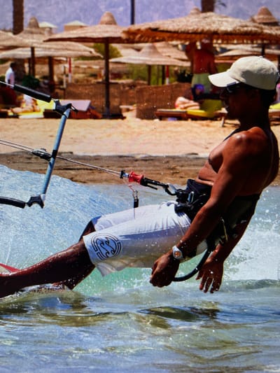 A man is kitesurfing in an area with shallow, clear water, likely a beach. The location could be Bacong, Philippines. The man is wearing a white cap, a black harness, and white shorts with a logo on the side. He is holding onto the kite control bar, and water is splashing around him. In the background, there is a sandy beach with beach umbrellas and possible other people.