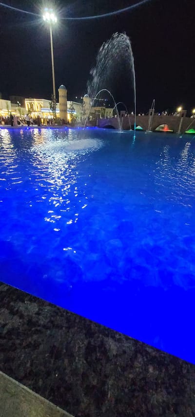 This is a nighttime outdoor photograph taken in the city of Shakhrisabz, Uzbekistan, specifically at the Nadir Divan-Beghi Madrasah complex. A large, illuminated pool of water dominates the foreground, glowing with an intense blue light from underwater fixtures. The water's surface reflects the light, creating shimmering patterns. In the background, a large fountain shoots water high into the air, illuminated by the surrounding lights.
Behind the fountain, a bridge with arched openings spans across a body of water, with people gathered along its edge. To the left of the fountain, the distinctive minarets and facade of the Nadir Divan-Beghi Madrasah are visible. The building is lit by warm, yellow lights, highlighting its architectural details. A tall, modern light pole with multiple floodlights stands prominently near the madrasah, casting bright white light. Several trees are scattered around the area, and many people are walking around, suggesting a public gathering or event. The overall atmosphere is lively and festive, with the combination of water features, illuminated architecture, and people creating a dynamic nighttime scene.