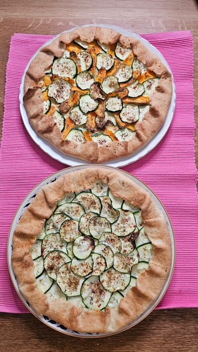 Two rustic tarts are presented on a wooden surface, each resting on a white plate. Both tarts feature a golden-brown, folded crust cradling a vibrant filling. The top tart displays a colorful medley of thinly sliced zucchini, orange squash, and possibly some other vegetables, seasoned with herbs. The bottom tart is primarily filled with overlapping slices of zucchini, also seasoned with herbs. Both tarts are placed on a bright pink placemat. The background shows a textured wooden surface. There are no people or animals present in the image, nor any clues indicating a specific location in Boussu-lez-Walcourt, Belgium.